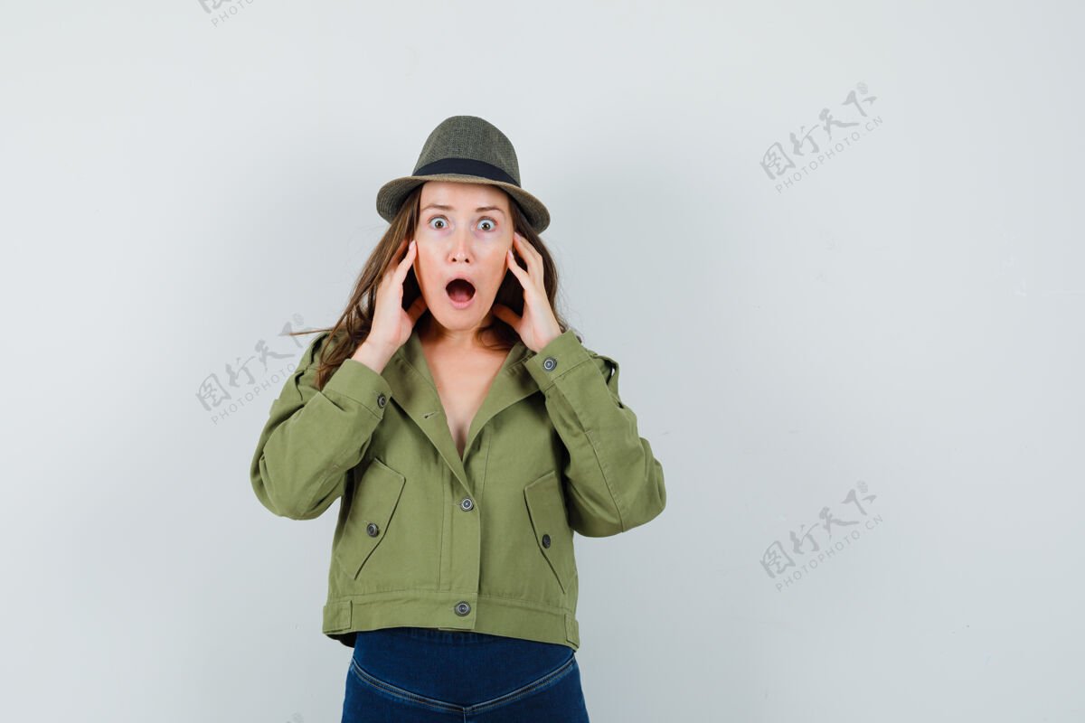 黑发穿着夹克 裤子 帽子的年轻女士手放在脸颊上 一边喊着 一边看起来很害怕拿着一起女性