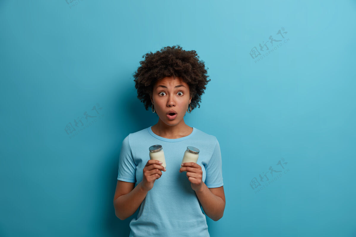 肖像吓得情绪激动的黑皮肤女人吓得直喘气 下巴一直耷拉着 手里拿着两杯酸奶有健康的营养坚持健康的饮食穿着休闲的蓝色t恤人 情绪 生活方式反应恐惧容器