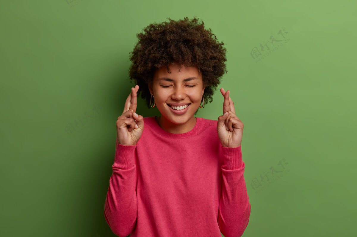 欲望快乐的非洲裔美国妇女的画像等待奇迹 祈祷和希望梦想成真 交叉手指祝好运 闭上眼睛 笑容满面 穿着粉红色的套头衫 隔离在绿色的墙上请欢乐微笑