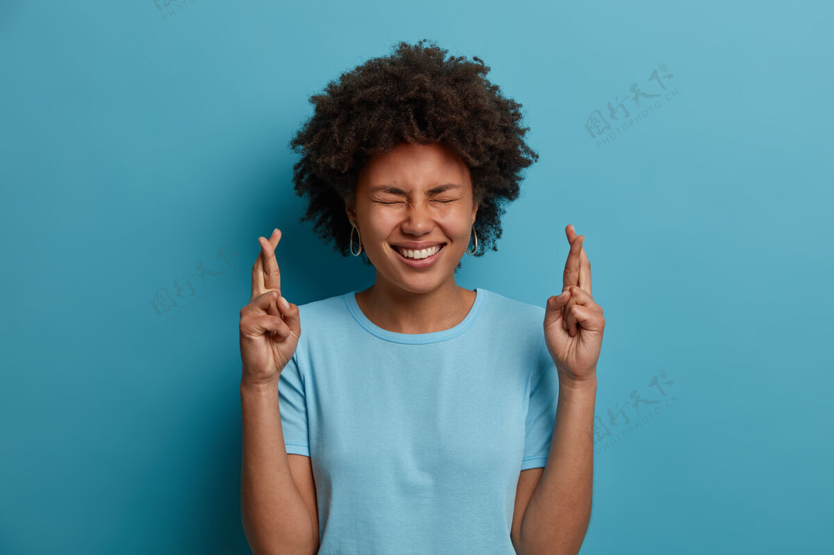 年轻积极的黑皮肤女人 非洲发型 为好运交叉手指 闭上眼睛 笑容灿烂 相信梦想成真 穿着休闲t恤 蓝色背景上孤立积极肖像发型