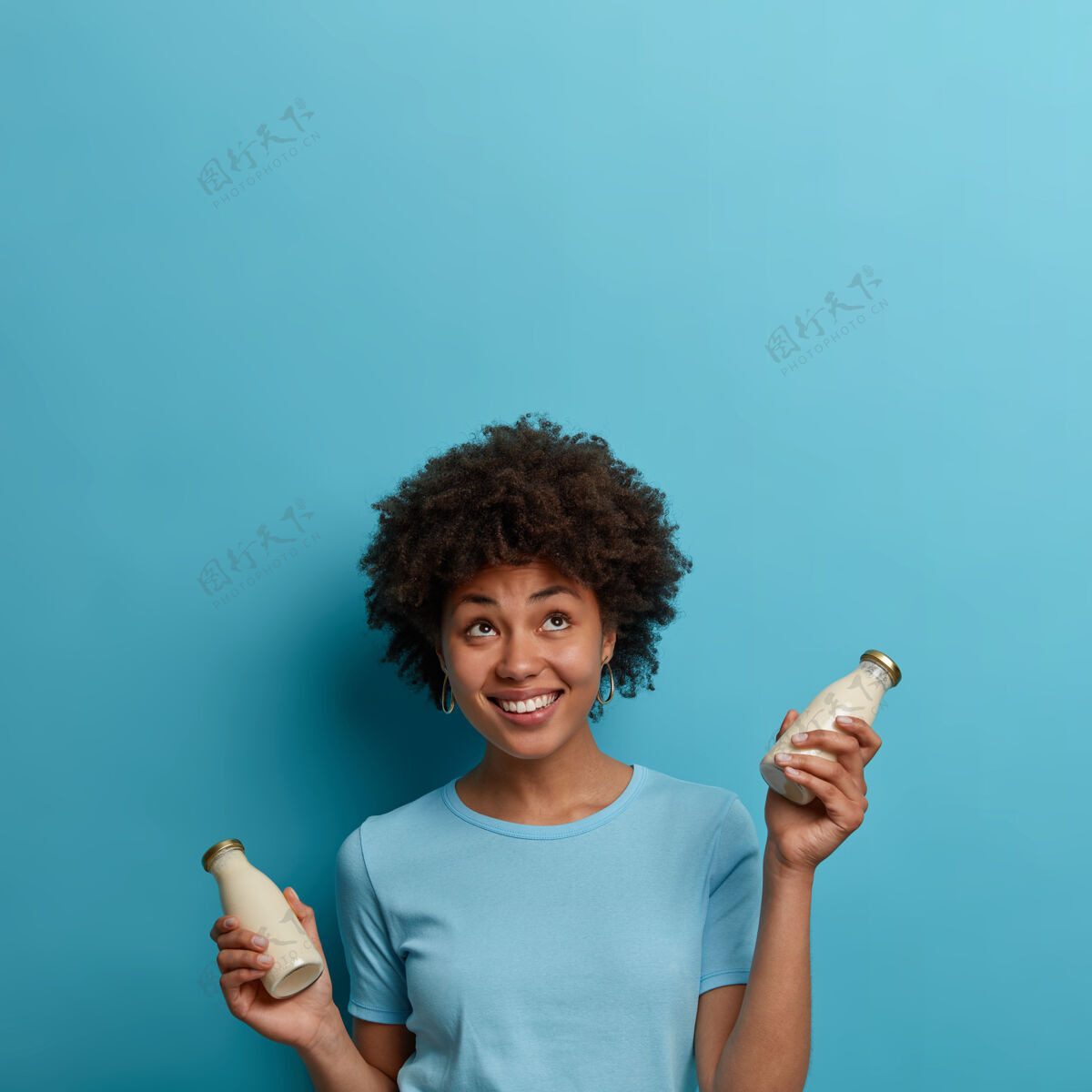 牛奶快乐的民族卷发女人喝无乳糖饮料 拿着一瓶杏仁或椰子奶 向上看 积极微笑 隔着蓝色的墙壁 复制空间为您的信息年轻提神奶瓶