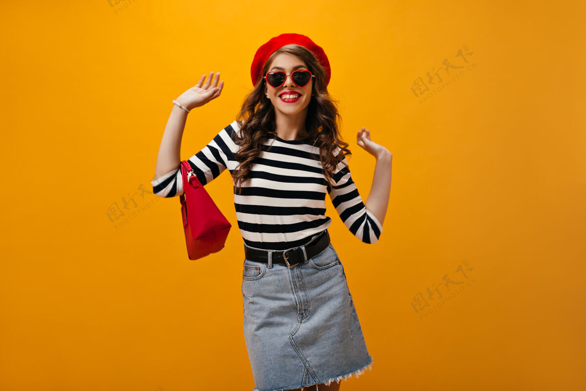 肖像戴着红色帽子和衬衫的漂亮女士在橙色背景上摆姿势带着包穿着条纹衬衫和时髦裙子的可爱女士微笑着成人休闲帽子