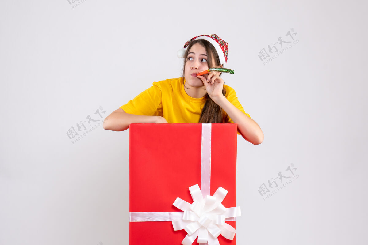 人前视图好奇的女孩 戴着圣诞帽 站在圣诞礼物后面 用隔音器快乐使用爱国