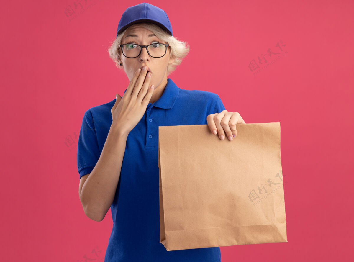 包装身穿蓝色制服 头戴眼镜 手拿纸包的年轻送货妇女被震惊 用手捂住嘴巴 翻过粉红色的墙壁年轻穿制服