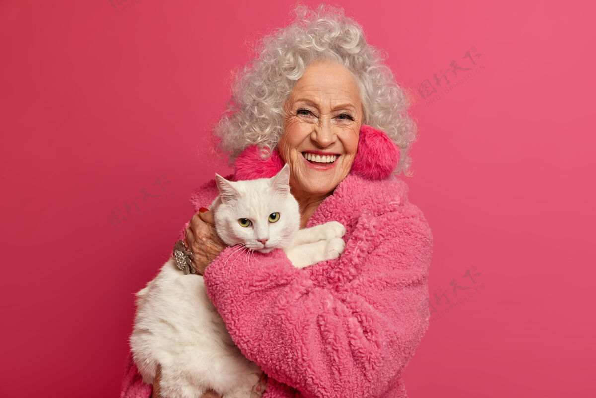 养老金满脸皱纹的祖母手牵白猫 大流行期间呆在家里 戴着蓬松的耳环 穿着柔软的长袍 准备喂宠物 隔离在粉红色的墙上女人领养老金主人朋友猫