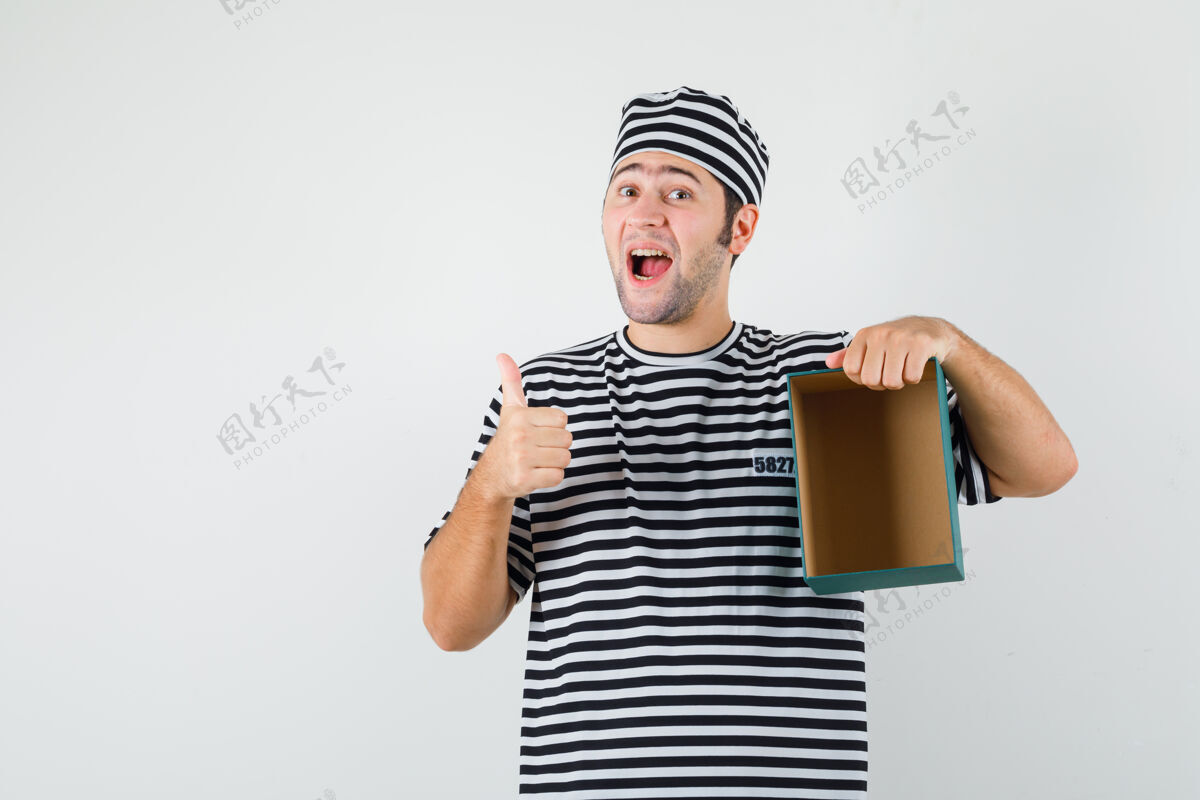 男人男青年穿着t恤 戴着帽子拿着空礼盒 竖起大拇指 看上去很开心 前视图拇指帽子时尚