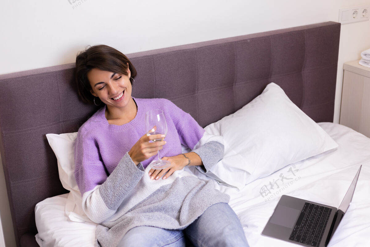 互联网快乐的女人穿着暖和的套头衫 躺在床上 一个人喝着一杯葡萄酒 休息着看喜剧电影 笑着笑着房间乐趣笑