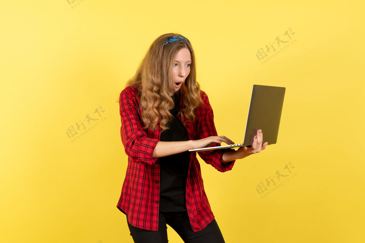 年轻女性正面图穿着红色格子衬衫的年轻女性在黄色背景上使用笔记本电脑女人人类情感模型时尚女孩人衬衫漂亮