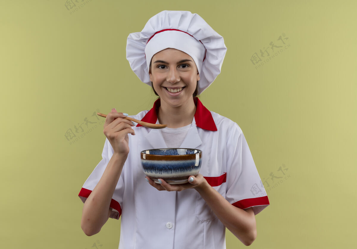 绿色身着厨师制服的年轻微笑的白人厨师女孩拿着碗和勺子放在绿色的空间里空间烹饪厨师