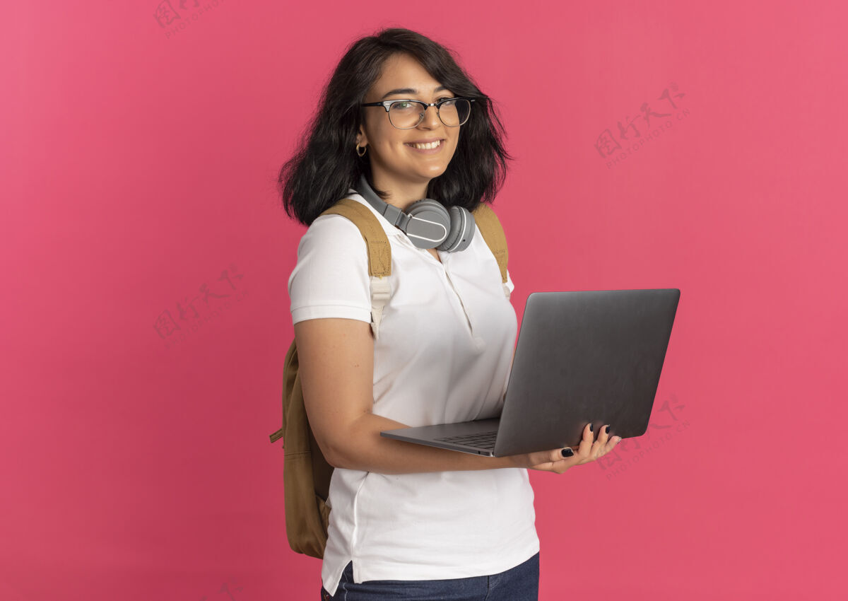 笔记本电脑年轻的白人女学生 面带微笑 脖子上戴着耳机 戴着眼镜 背着书包 粉色的笔记本电脑上有复印空间穿女学生年轻