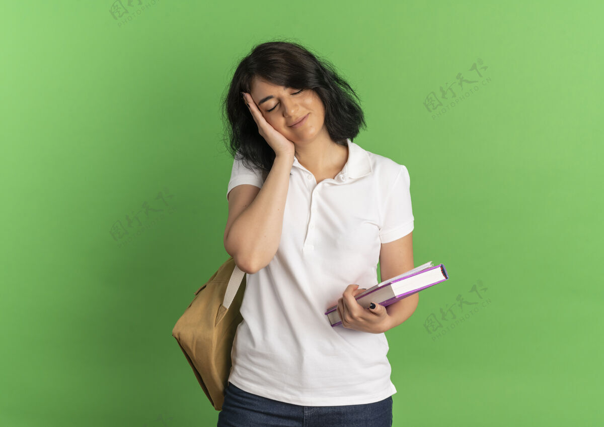 抱年轻可爱的高加索女学生戴着书包 手放在脸上 手里拿着书 放在绿色的书架上 留着复印空间穿请空间