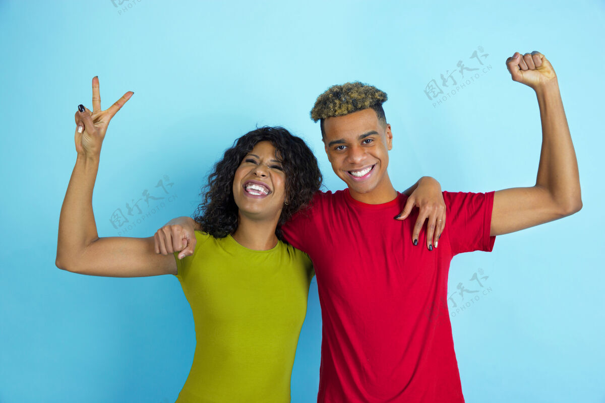 朋友摆出胜利者的样子 笑着年轻的情绪化的非裔美国人在蓝色的墙上穿着五颜六色的衣服情绪年轻笑