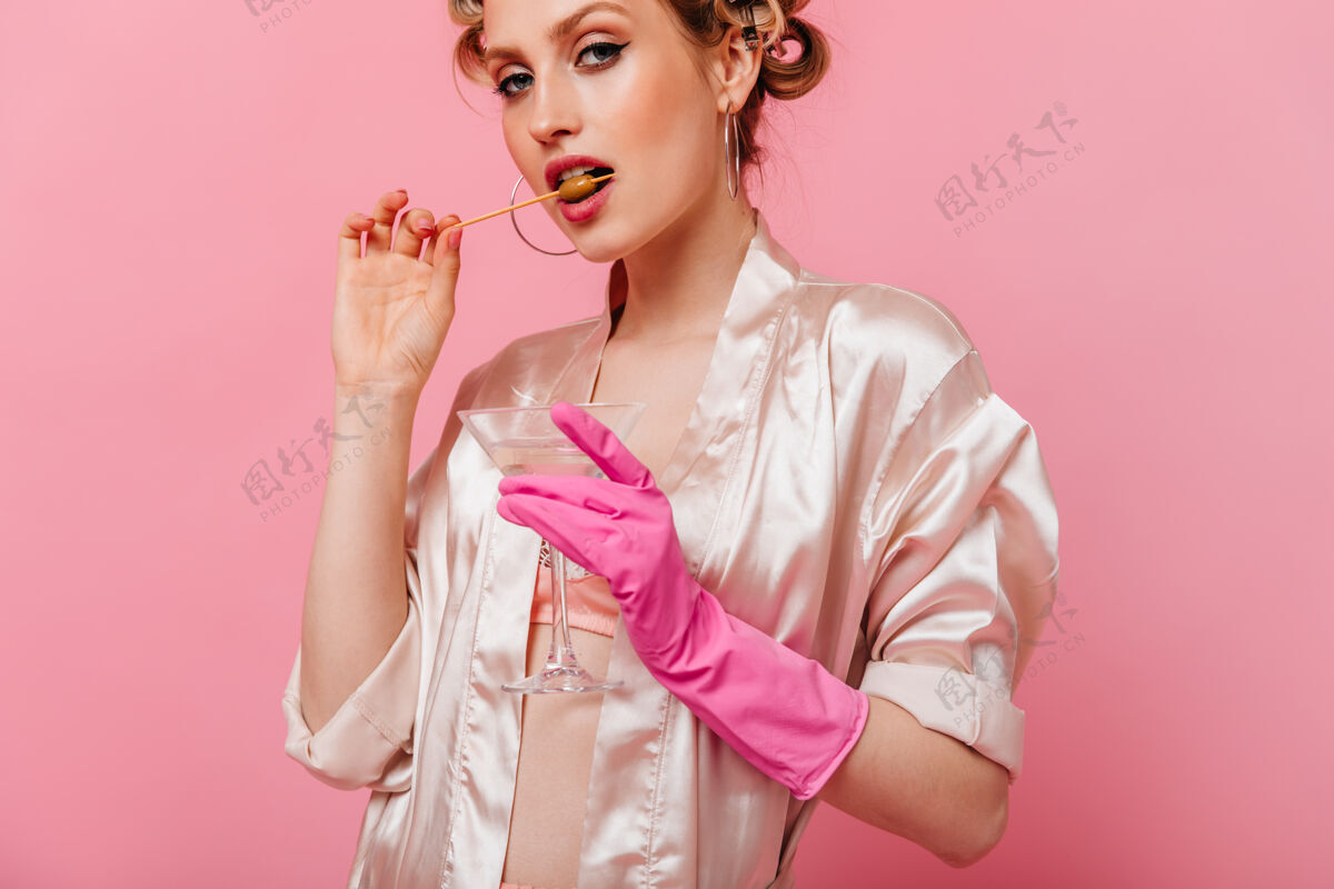 美丽粉红色墙上咬着橄榄 拿着马提尼杯的女人的画像享受浴袍成人