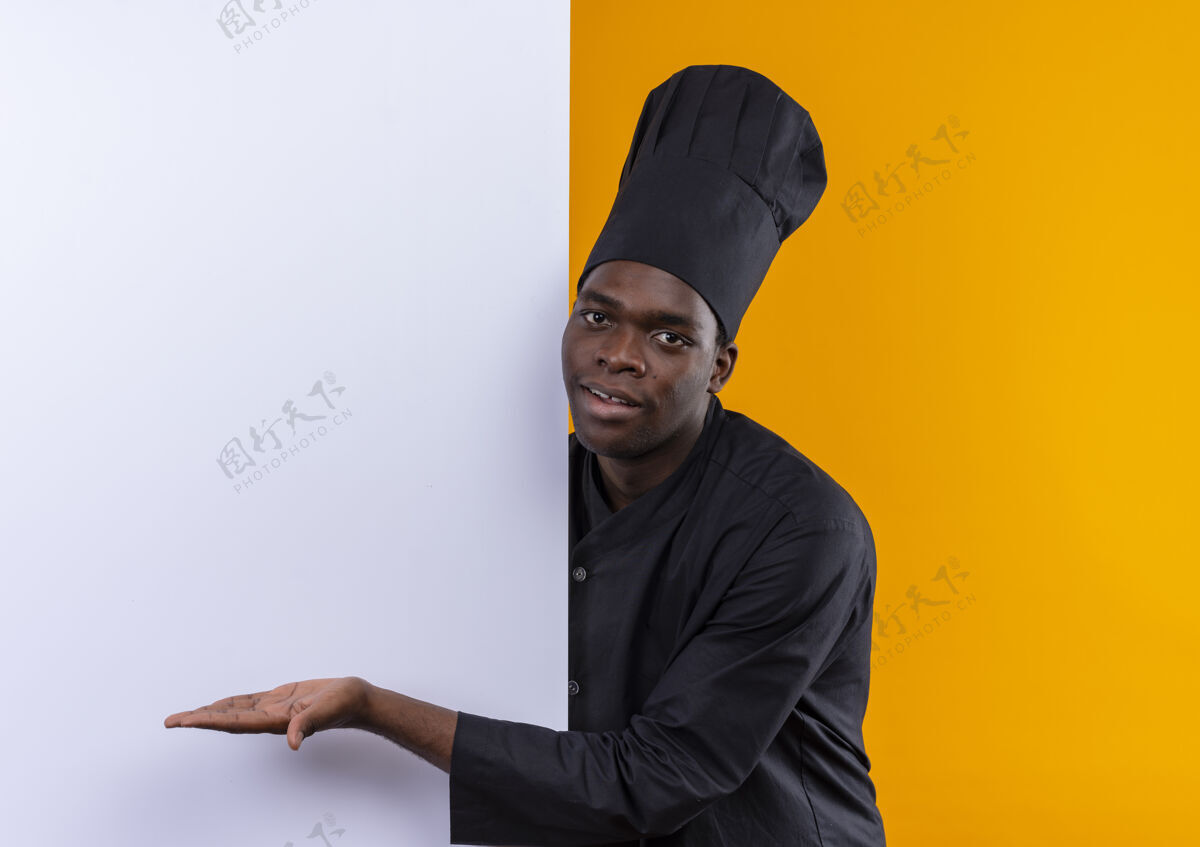 请身着厨师制服的年轻美籍黑人厨师站在身后 用手指着橙色的白色墙壁 上面留有复印空间厨师空间复制