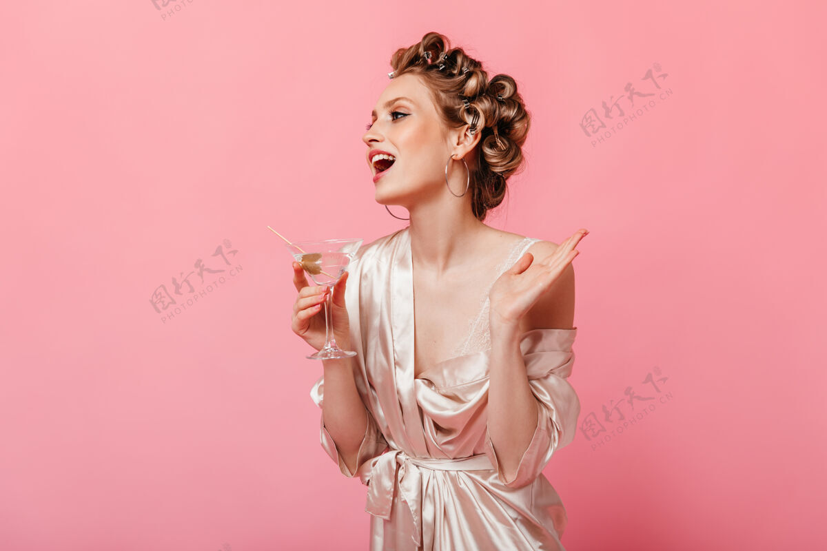 欢呼戴着卷发器和浴袍的女士笑着 在隔离墙上摆着马提尼杯的姿势女性杯子丝绸