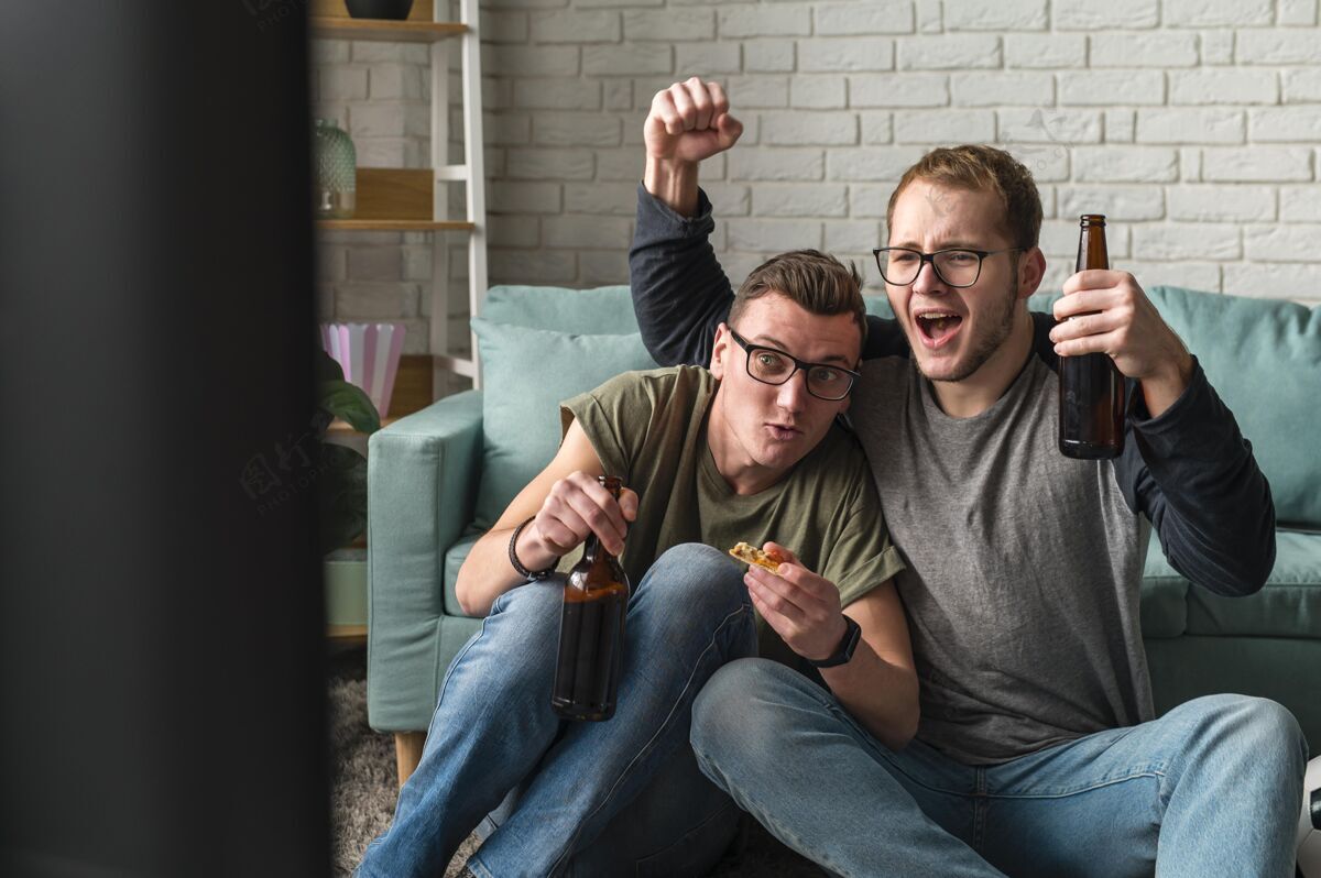 欢呼两个快乐的男性朋友在电视上看体育节目和喝啤酒的正面图一起伙伴男