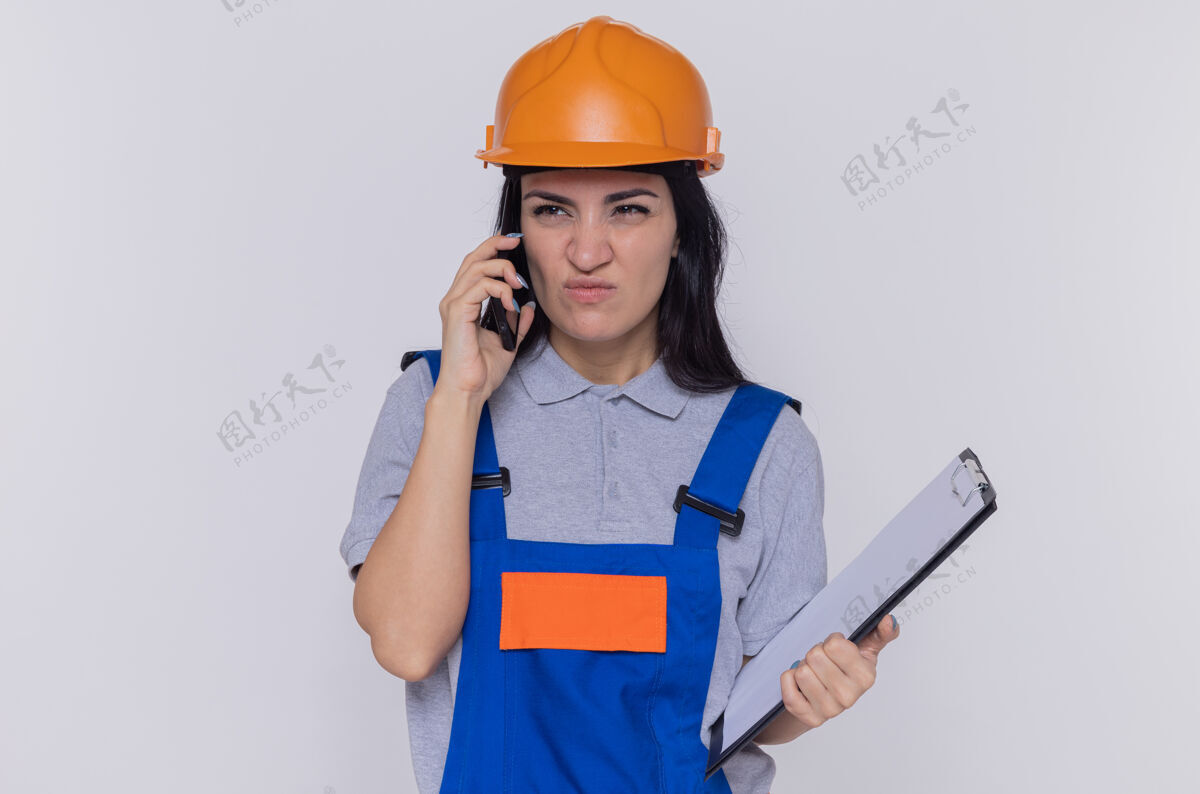 站着身穿建筑制服 戴着安全帽 拿着写字板的年轻建筑工人站在白色的墙上 一边用手机说话 一边显得很不高兴拿着年轻人女人