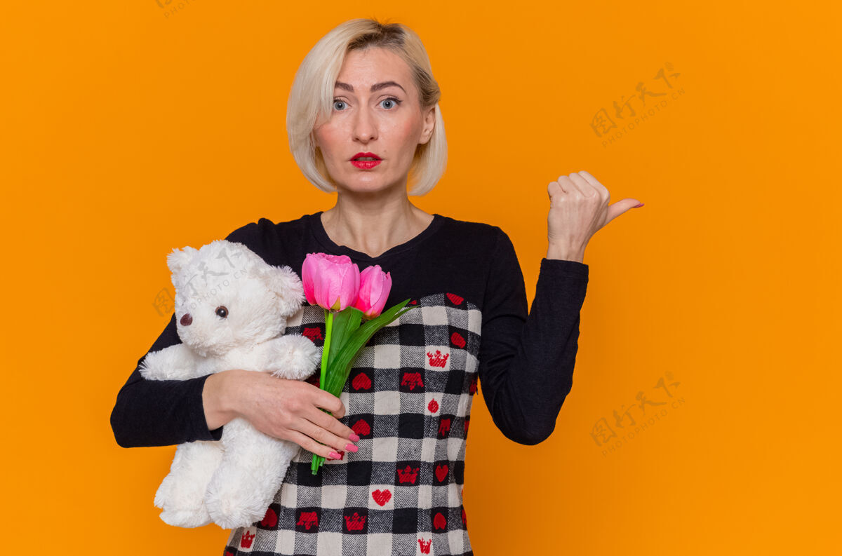 熊困惑的年轻女子穿着漂亮的衣服 手持郁金香和泰迪熊的花束作为礼物 用拇指指着身后站在橙色的墙上庆祝国际妇女节美丽女人国际