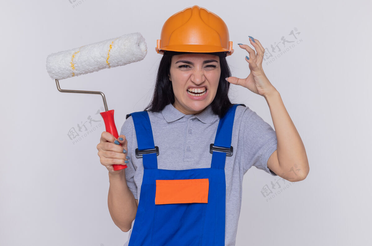 安全身穿施工制服 戴着安全帽的年轻建筑工人举着油漆辊 怒气冲冲地看着前面 她举起胳膊站在白色的墙上滚轴建筑女人