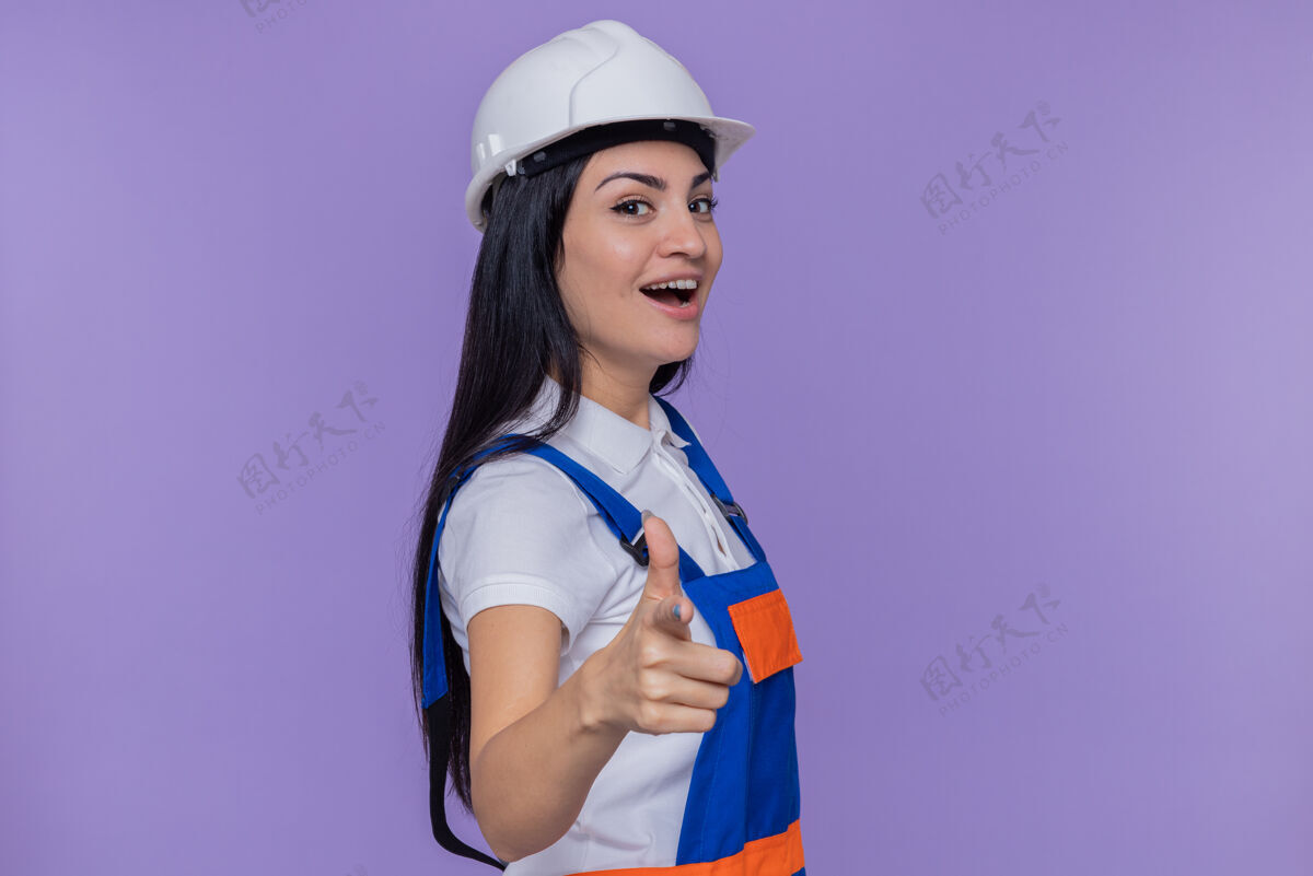 站立身穿施工制服 头戴安全帽的年轻建筑工人站在紫色的墙上 用食指指着前面 脸上带着羞涩的微笑年轻人微笑安全
