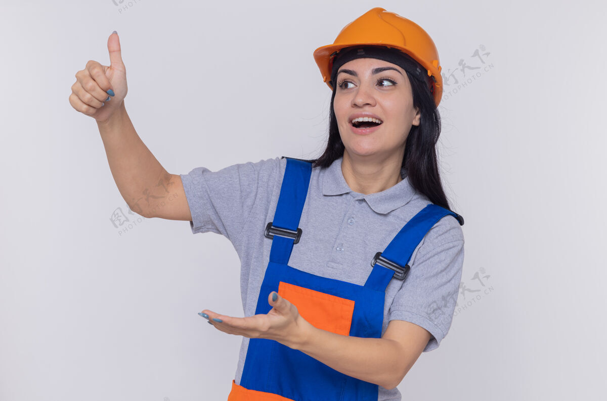 微笑身穿施工制服 头戴安全帽的年轻建筑工人一边微笑一边举起大拇指一边伸出手臂站在白墙上建筑建筑工人女人