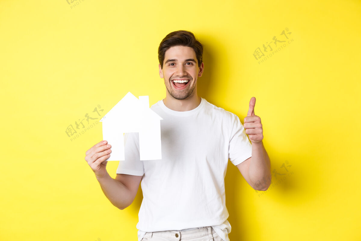 年轻房地产概念快乐的年轻人展示纸房子模型 竖起大拇指 推荐经纪人 站在黄色背景下模特男人站着