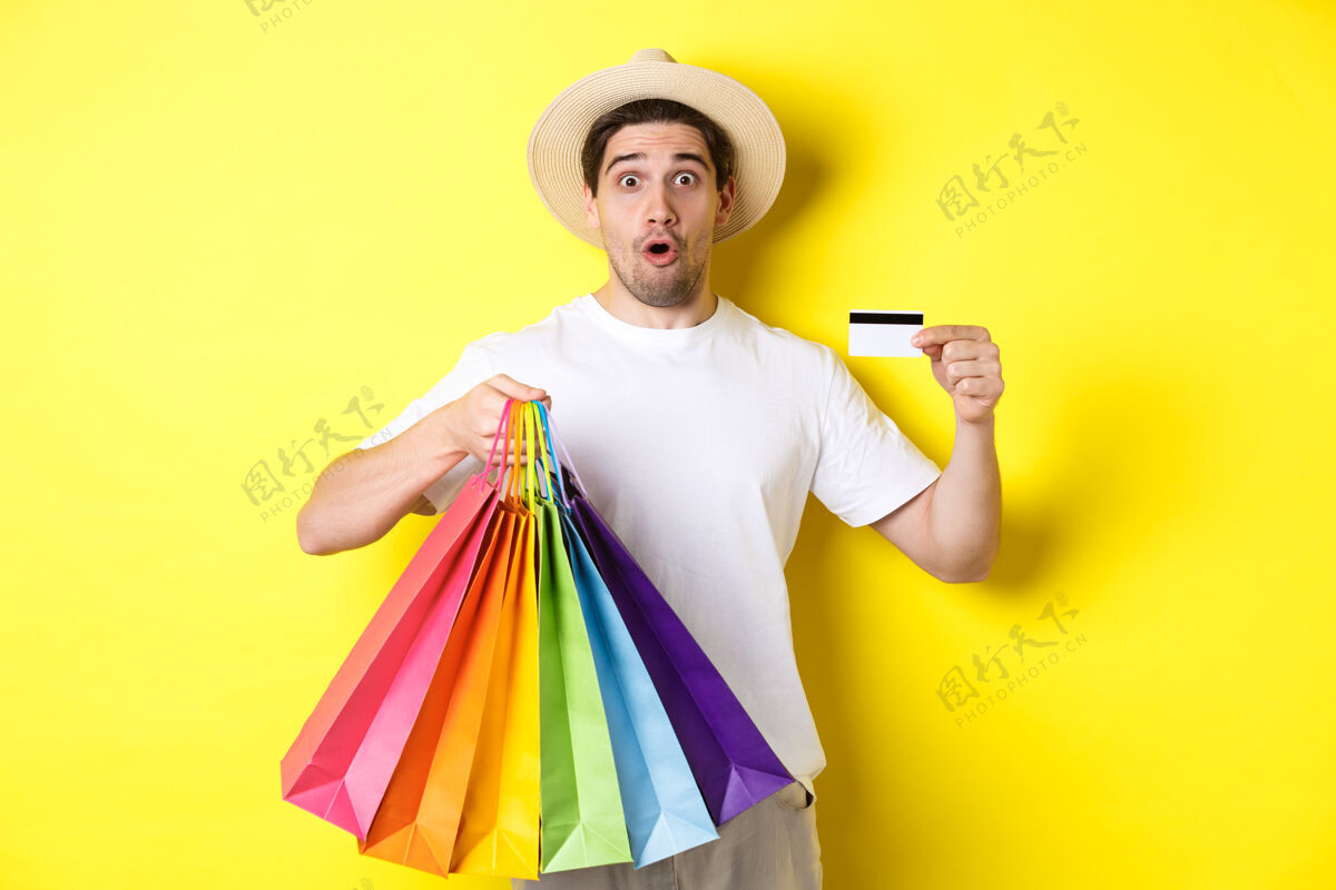 工作室令人印象深刻的男子展示购物袋与产品和信用卡 站在黄色背景帽子男人购物袋