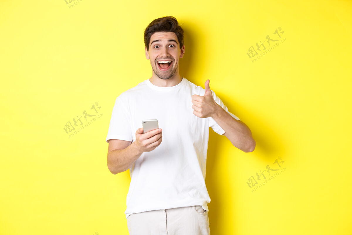 工作室快乐的微笑的男人拿着智能手机 竖起大拇指表示赞同 在网上推荐一些东西 站在黄色的背景上男人成人男人