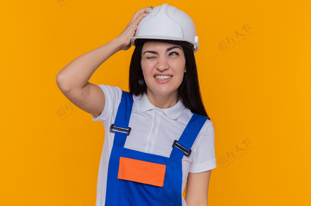 头身穿施工制服 头戴安全帽的年轻建筑妇女站在橙色的墙上 手举着头 脸上带着恼怒的表情头盔年轻建设者