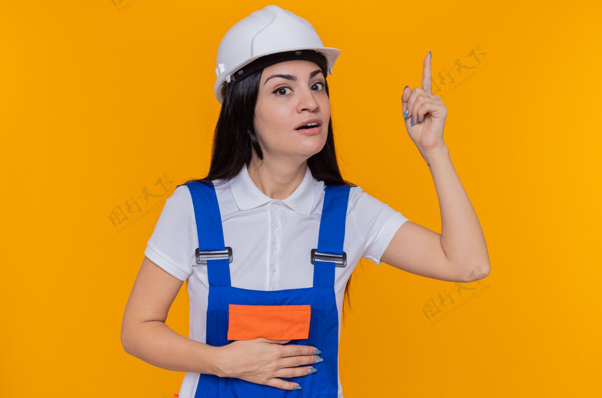 手指身穿施工制服 头戴安全帽的年轻建筑工人站在橙色的墙上 面带微笑地看着前面 脸上露出惊讶的表情 食指上有着伟大的想法聪明头盔伟大