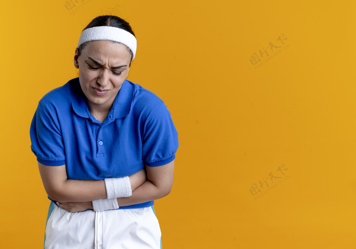 疼痛年轻疼痛的高加索运动女性戴着头带和腕带 腹部放在橘黄色的复制空间上年轻腹部持有