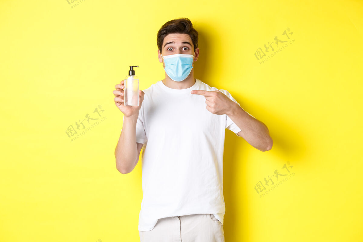 帅气covid-19的概念 检疫和生活方式兴奋的家伙戴着医用口罩 展示了很好的洗手液 指着防腐剂 站在黄色的背景上演播室时尚洗手液