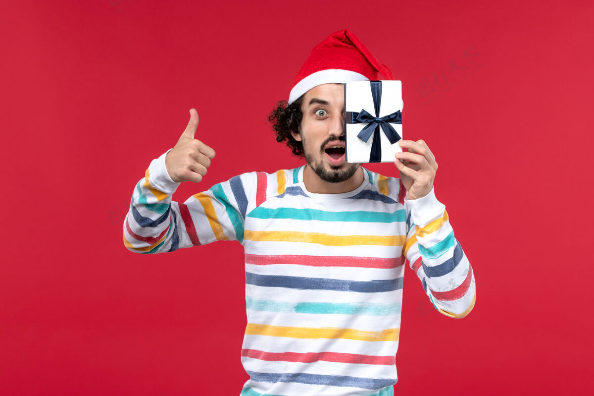 人正面图红墙上年轻男子手持小礼物过年情感红帽子微笑圣诞节