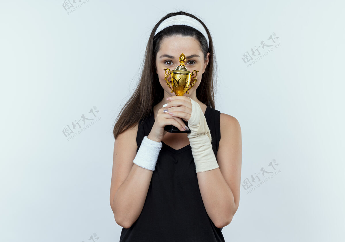 站着戴着头巾的年轻女健身者高高兴兴地举着她的奖杯站在白墙上 脸上洋溢着积极的笑容抱着年轻头带