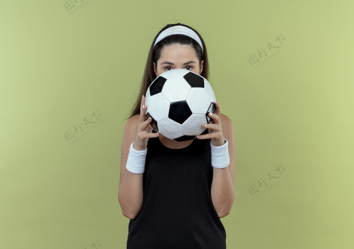 年轻戴着足球头巾的年轻健身女士把脸藏在球后 站在轻质墙上偷看轻女子球