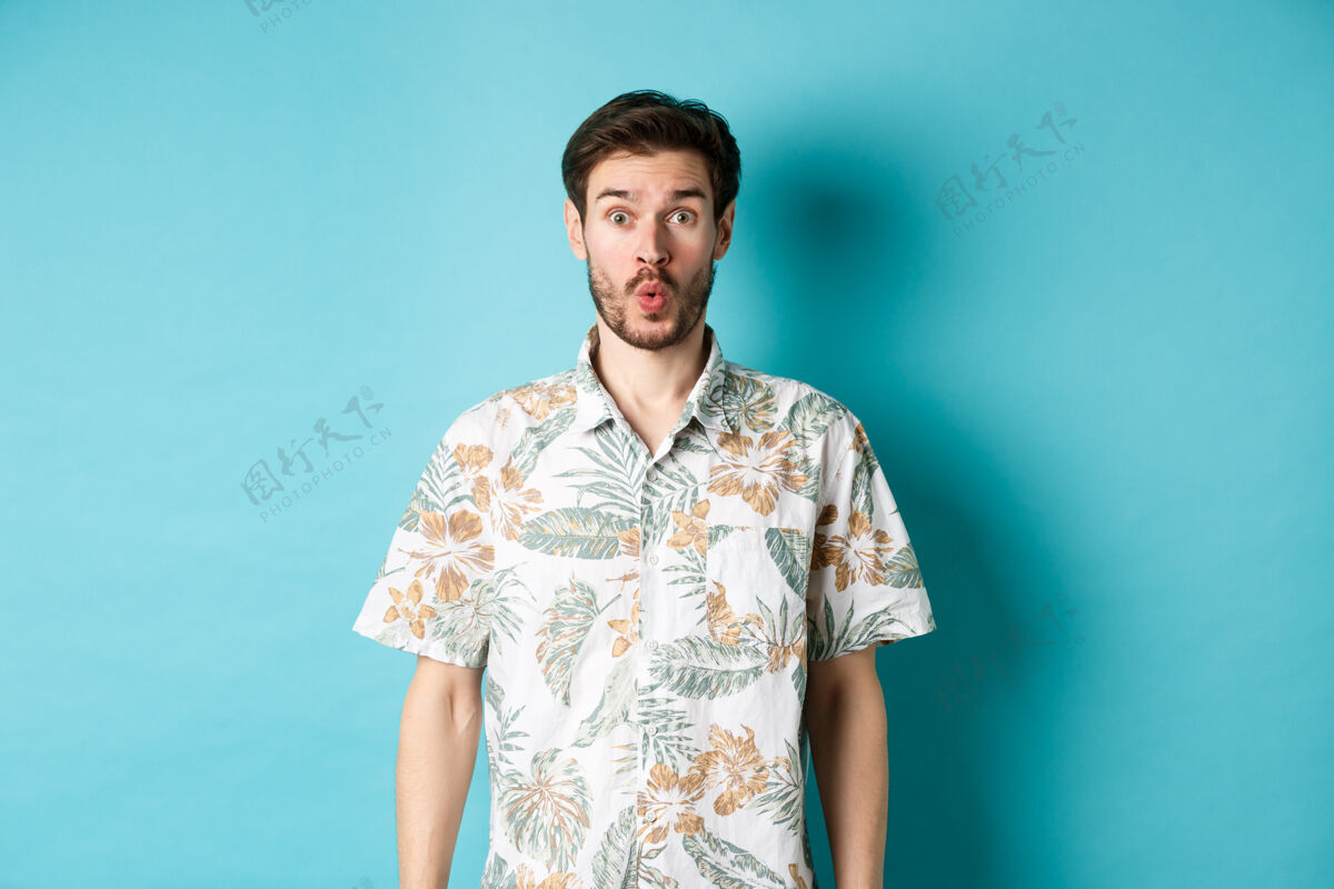室内暑假惊讶的游客说哇哦 盯着相机 查看真棒的宣传片 站在蓝色背景的夏威夷衬衫酷男人自信