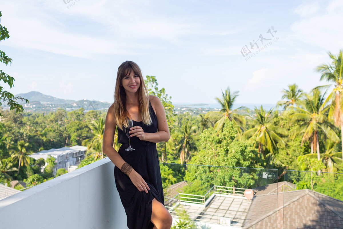 成人在热带阳台上 一个穿着黑色晚礼服 端着一杯葡萄酒的女人人女孩玻璃