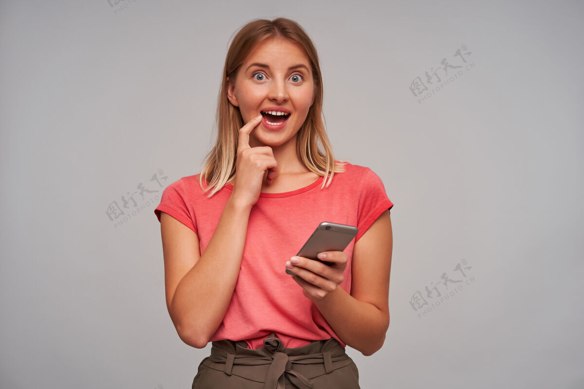 兴奋十几岁的女孩 一头金发 看上去很快乐穿着粉色t恤和棕色裙子拿着智能手机 摸着嘴角肖像电话女性