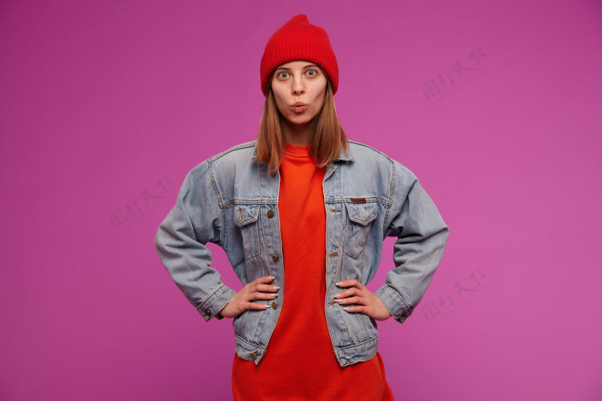 肖像少女 快乐的女人 深色长发穿着牛仔裤夹克 红色毛衣和帽子把手放在腰上 在紫色的墙上做亲吻的脸人面部学生