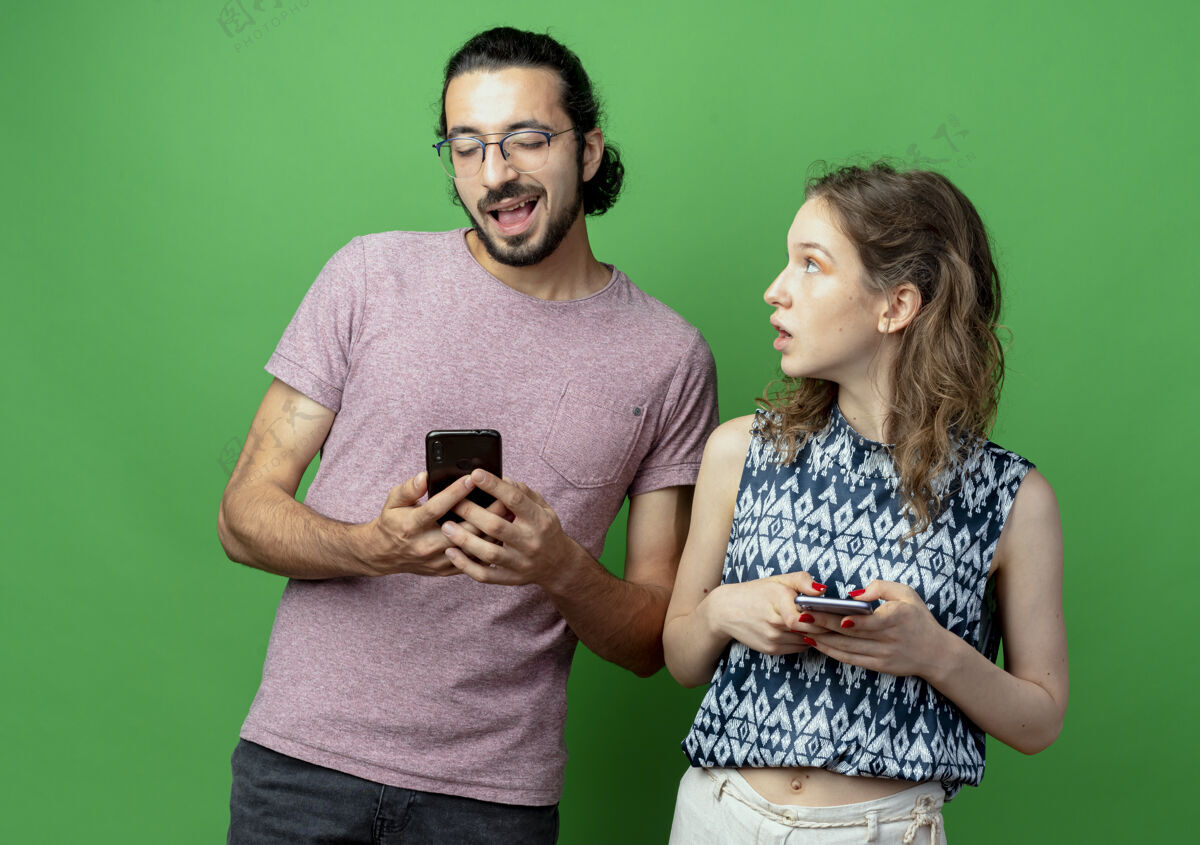 情侣一对年轻夫妇拿着智能手机 一个女人惊讶而困惑地看着自己的男朋友翻过绿墙男朋友惊喜年轻