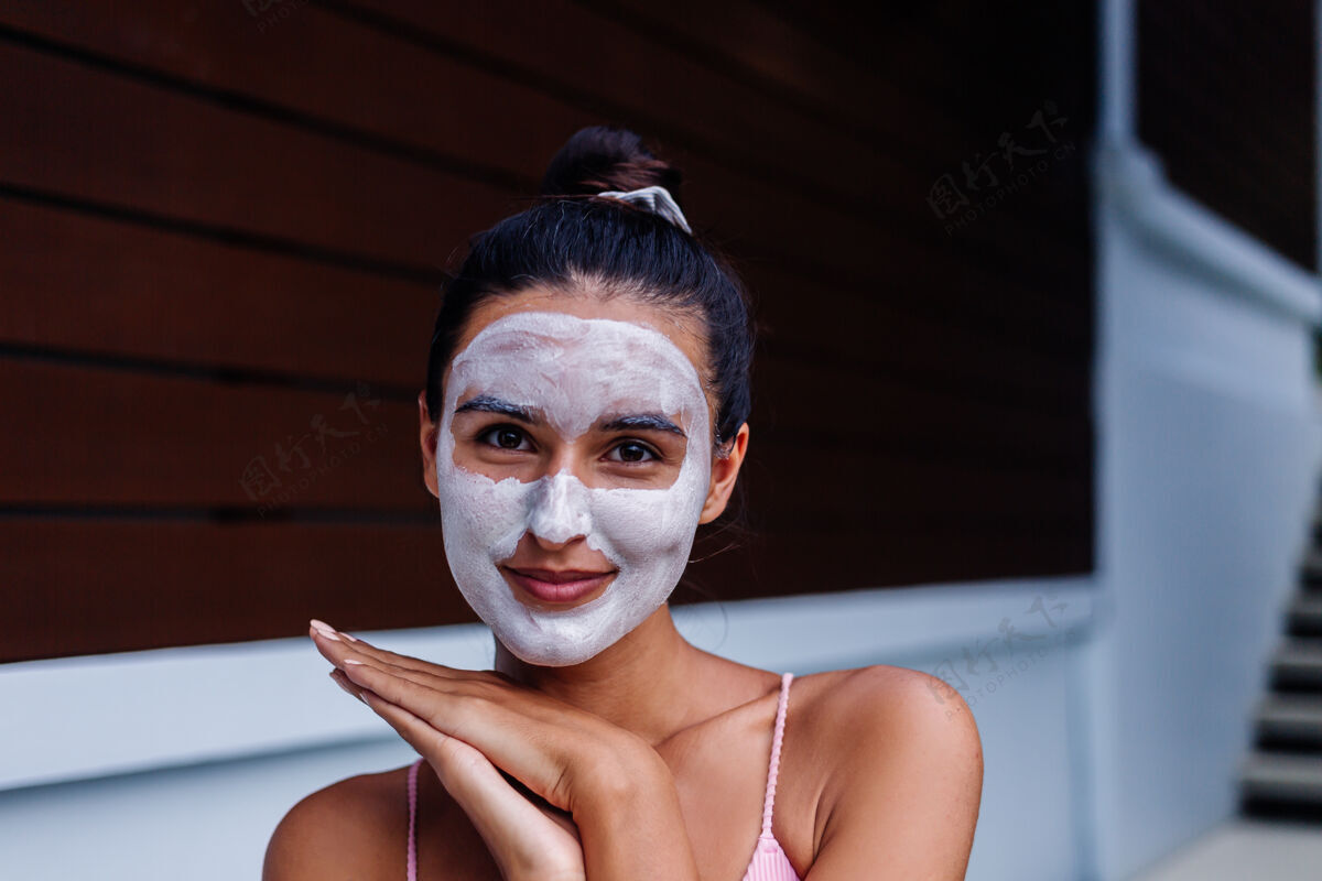 面部晒黑皮肤的户外写真美丽的高加索女子穿着比基尼在温泉浴场与白色脱皮面具在脸上更新自然清洁
