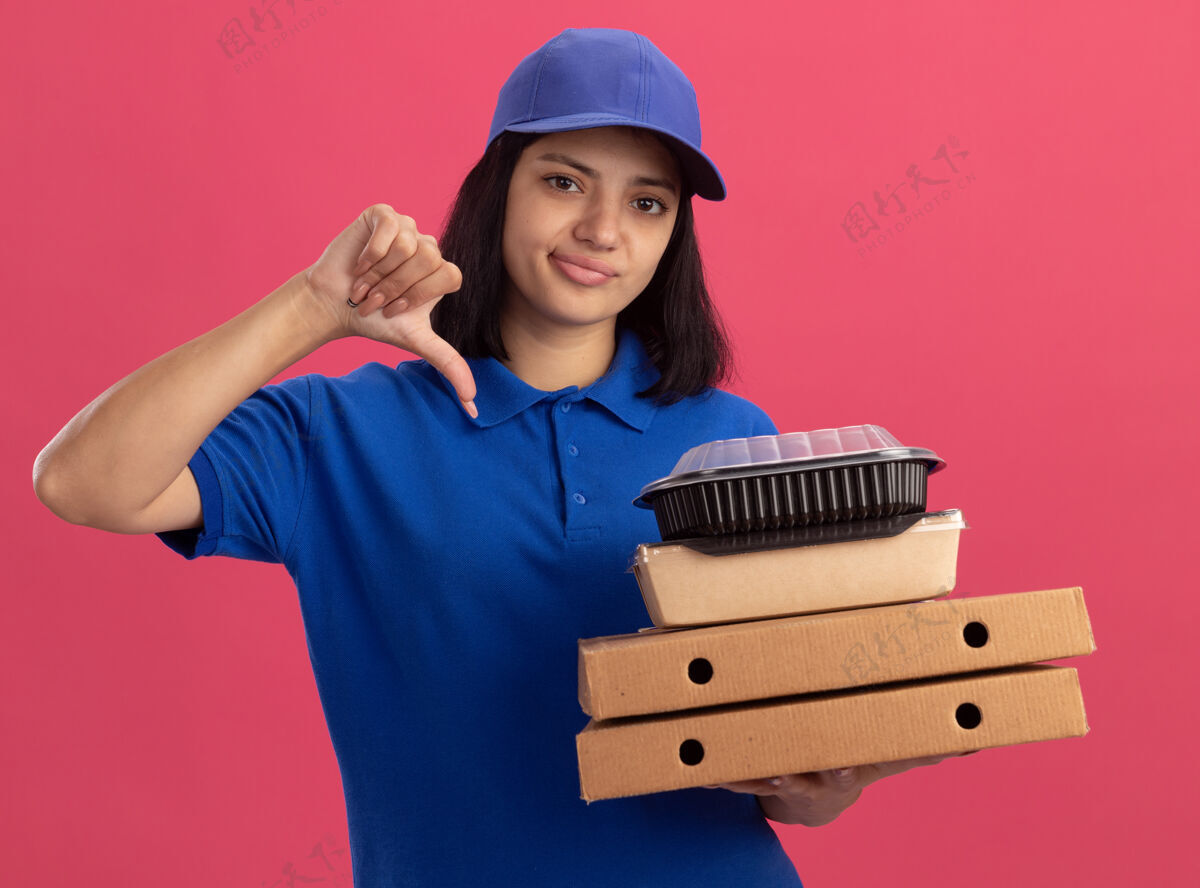 放下不满意的年轻送货员身穿蓝色制服 头戴鸭舌帽 手里拿着比萨饼盒和食品包 站在粉红色的墙上 看起来大拇指朝下帽子食物盒子