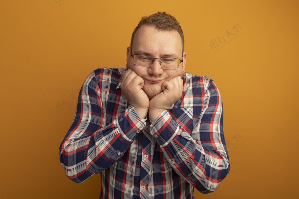 男人男人戴着眼镜 穿着格子衬衫 手贴着脸 开心地站在橘色的墙上 感受着积极的情绪欢呼眼镜站着