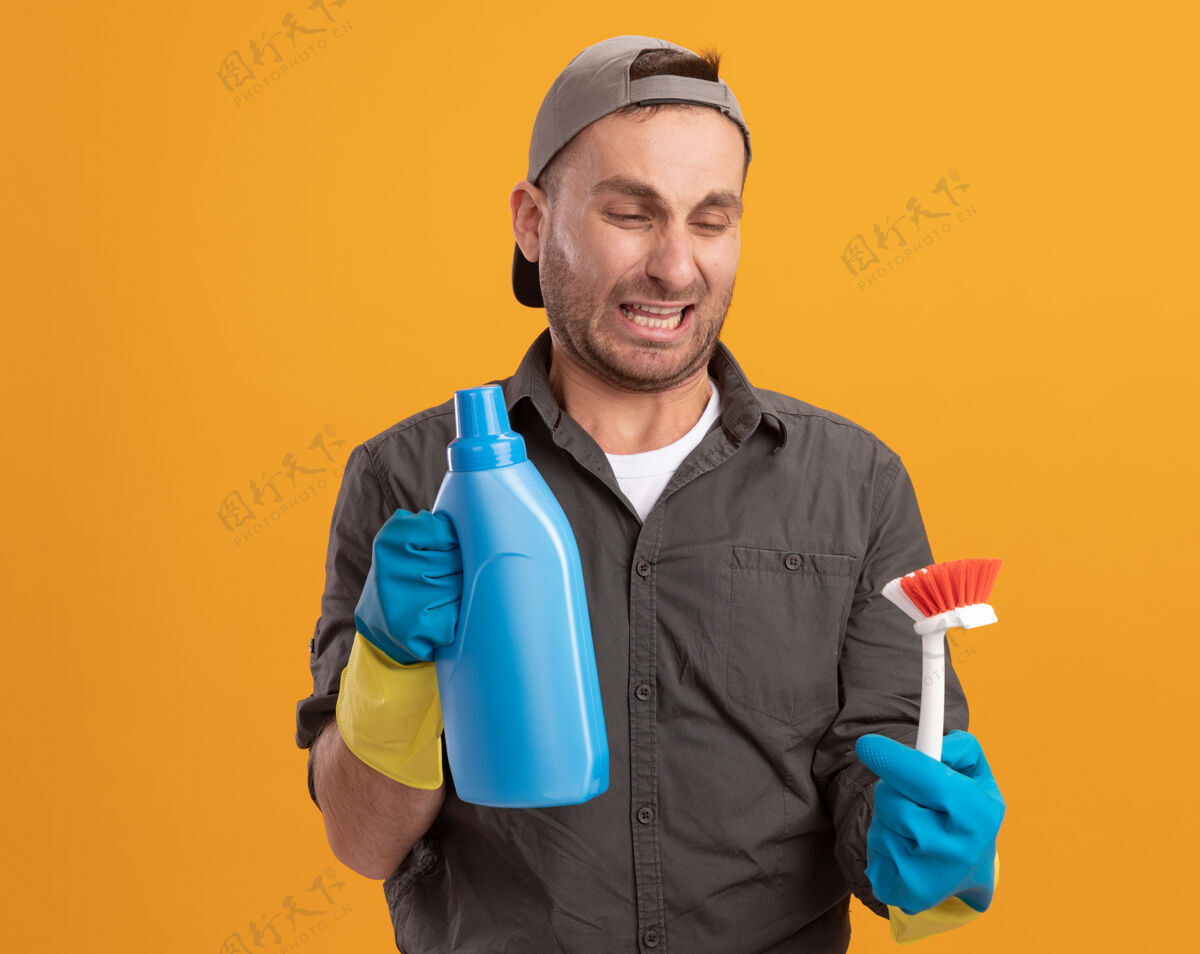 男人年轻的清洁工穿着便服 戴着橡胶手套 戴着帽子 手里拿着清洁刷和瓶子 手里拿着清洁用品 带着厌恶的表情看着刷子站在橙色的墙上瓶子站着年轻