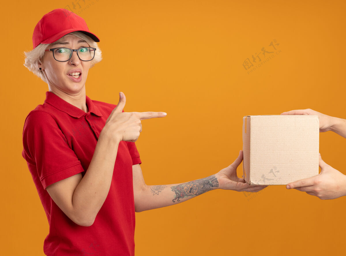 困惑身穿红色制服 戴着帽子的年轻送货员在收包裹时感到困惑 食指指着站在橙色墙上的盒子目录年轻接收