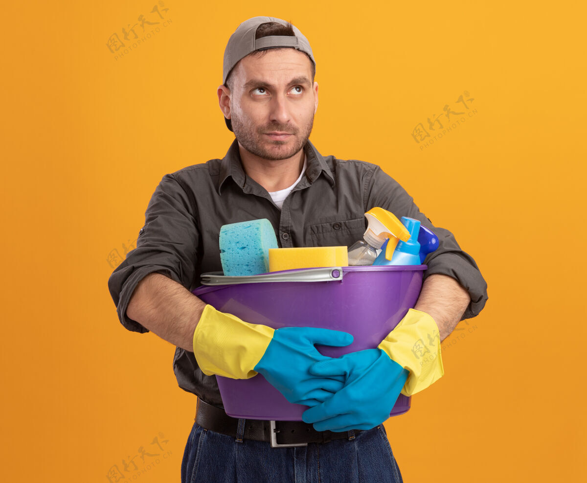男人年轻人穿着休闲服 戴着橡胶手套 手里拿着水桶 手里拿着清洁工具 站在橘红色的墙上 抬头看着困惑和不快工具桶年轻