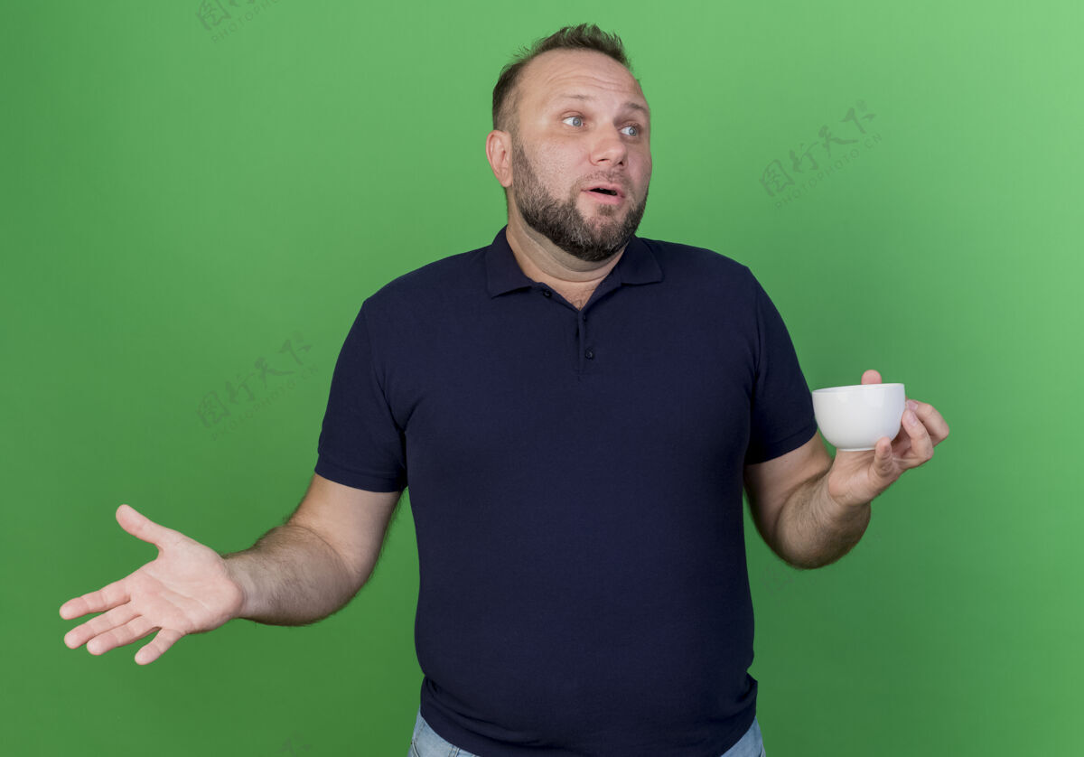 男人令人印象深刻的成年斯拉夫男子拿着杯子 显示空的手看着隔离在绿色墙上的一面看成人杯子
