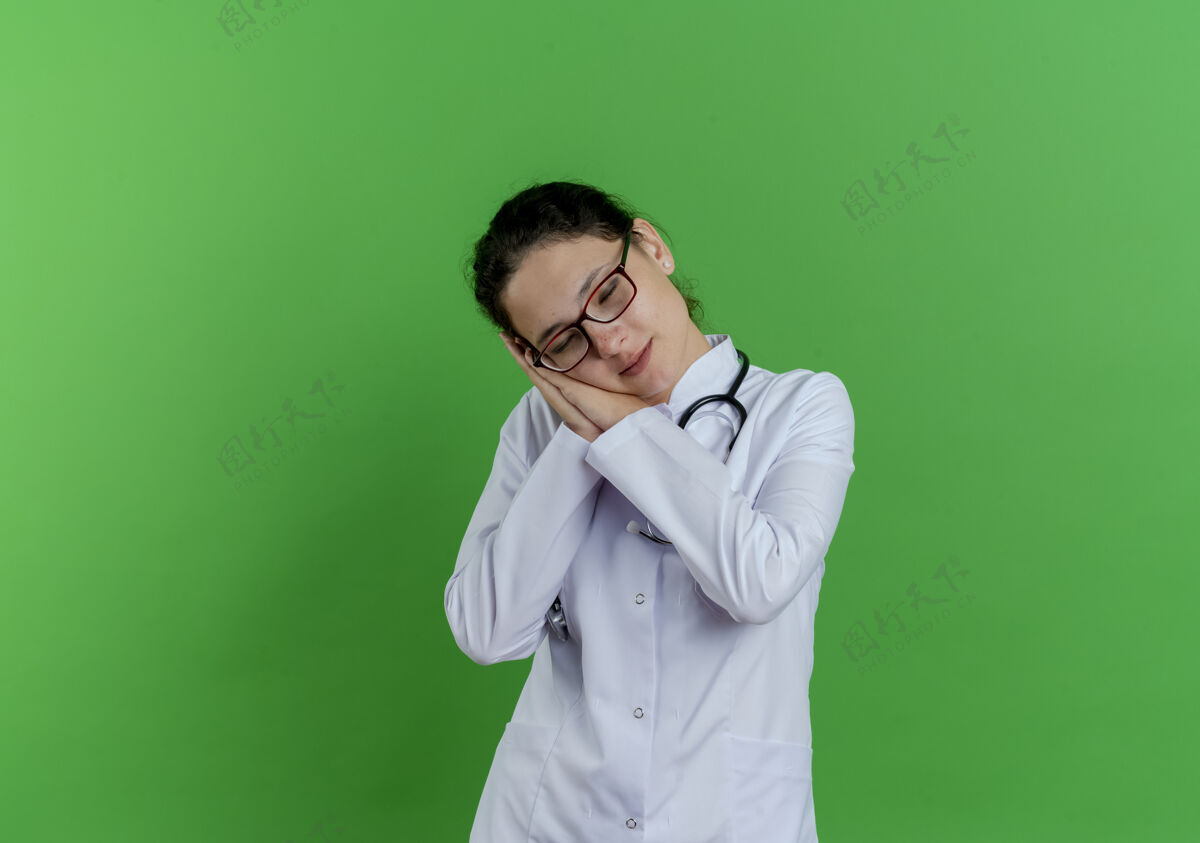 穿疲惫的年轻女医生 穿着医用长袍 戴着听诊器和眼镜 做着闭着眼睛睡觉的姿势 隔离在绿色的墙上 留着复印空间听诊器疲倦医疗