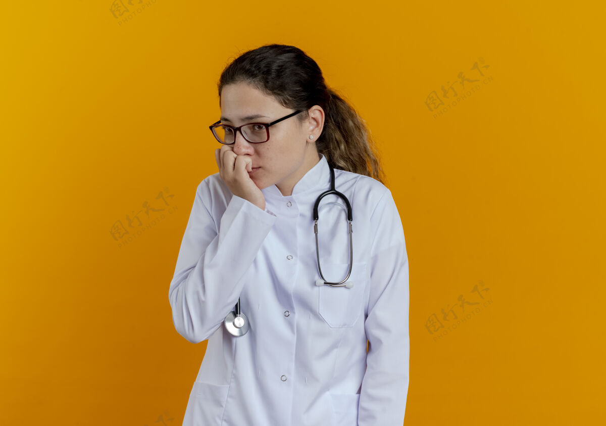 长袍看着身边忧心忡忡的年轻女医生穿着医用长袍 戴着眼镜的听诊器咬着橘黄色墙上孤立的钉子看指甲壁板