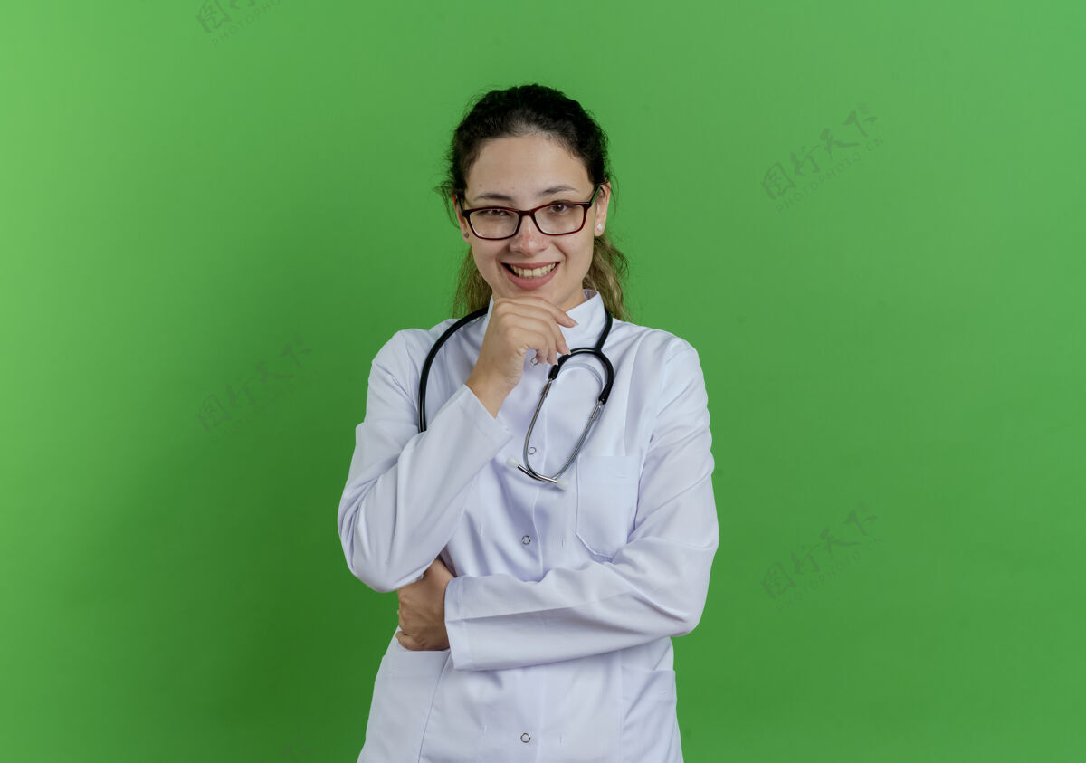 眼镜面带微笑的年轻女医生 穿着医用长袍 戴着听诊器 戴着眼镜 触摸着隔离在绿色墙壁上的下巴 还有复印空间下巴触摸医疗